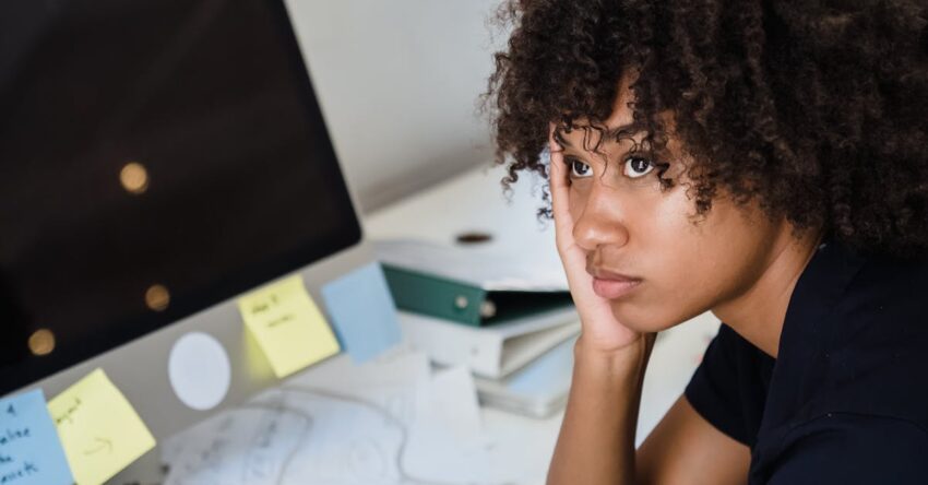 A woman looks tired while sitting at her work desk surrounded by computers and paperwork.