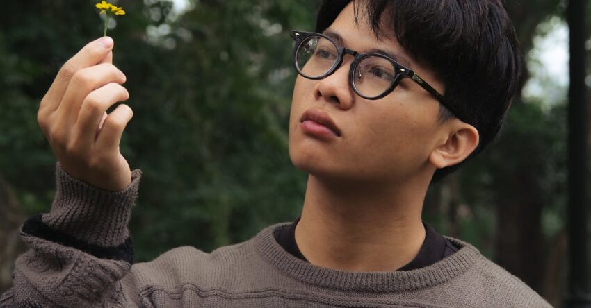 Young man wearing a patterned sweater holding a yellow flower outdoors.