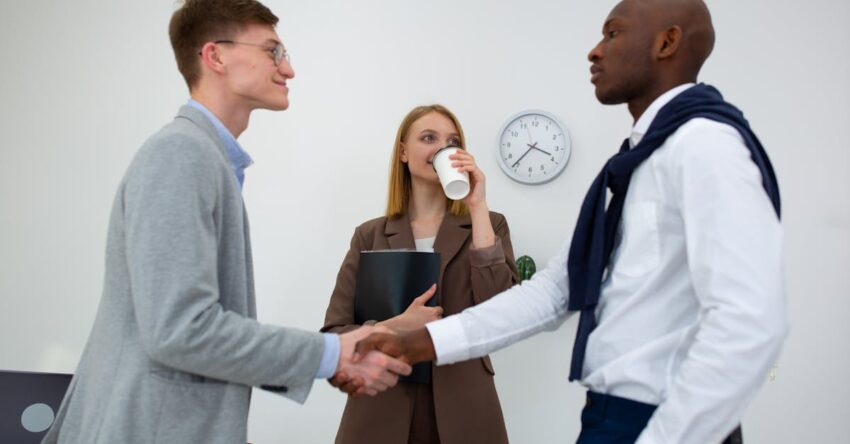 Professional meeting with two men shaking hands and a woman with a coffee cup in an office.