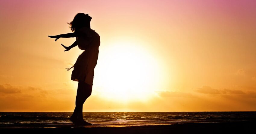 Silhouette of a woman at the beach with arms outstretched against a vibrant sunset backdrop.