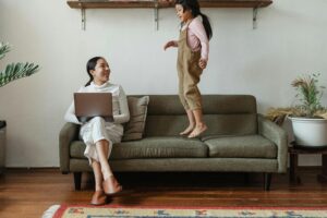 Smiling Asian mother in casual wear working on laptop while cute daughter jumping on couch in cozy living room