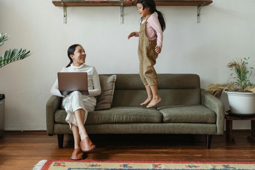 Smiling Asian mother in casual wear working on laptop while cute daughter jumping on couch in cozy living room